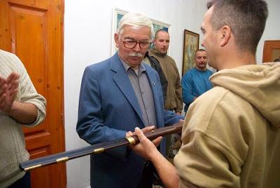The picture shows Győrkös, a 70-year-old man with gray hair and a mustache, receiving an antique rifle from another man.
