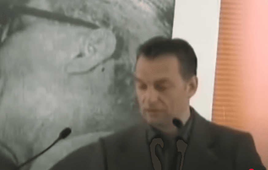 The picture shows Viktor Orbán giving a speech at a party event. The background shows Leonid Brezhnev and Erich Honecker engaging in a fraternal kiss.